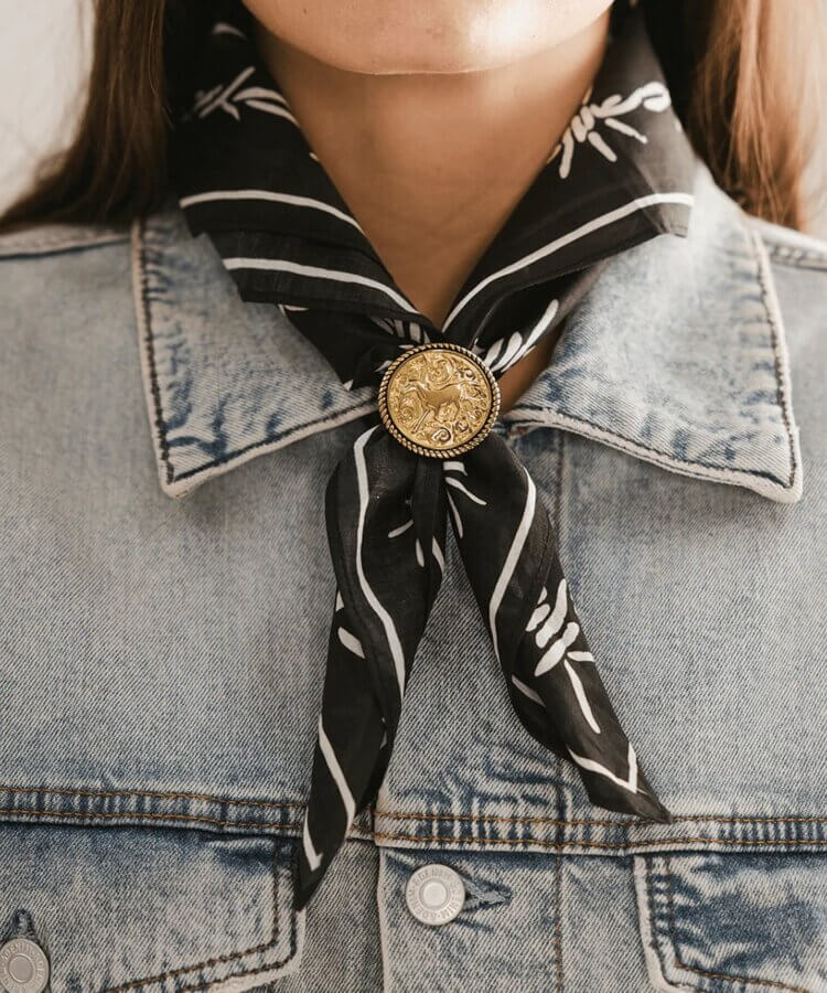It's the Year of the Brooch! Here's How to Style Them - 4 A person wearing a light denim jacket and a black patterned scarf tied at the neck with a large gold decorative pin, showcasing their love for stylish brooches.
