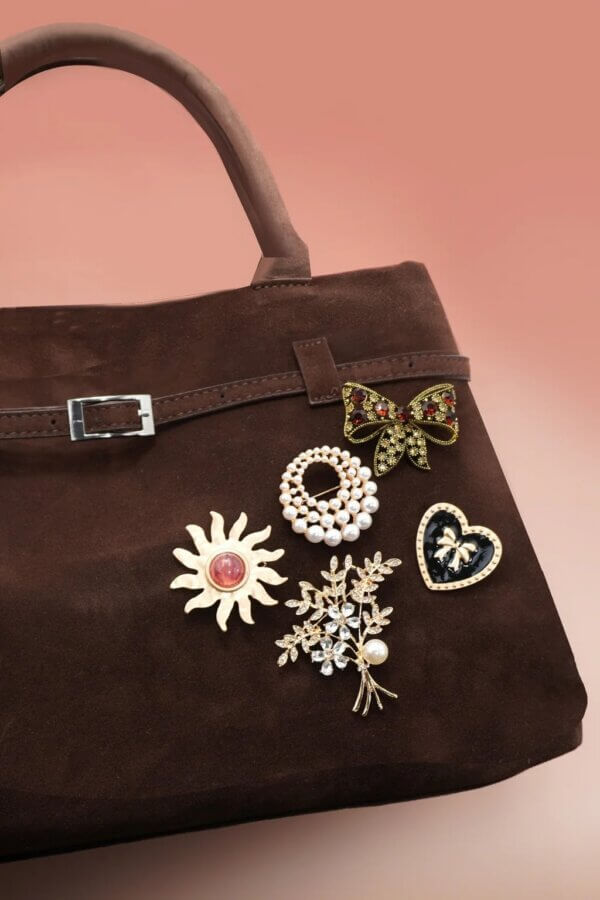 It's the Year of the Brooch! Here's How to Style Them - 15 A brown suede handbag adorned with five unique broochesβa bow, sun, pearl circle, floral branch, and heartβstands out against a pink gradient background.