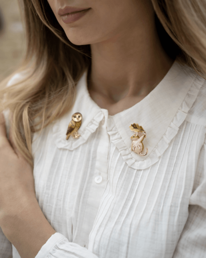 It's the Year of the Brooch! Here's How to Style Them - 12 A woman in a white blouse with pleated details wears two charming broochesβone shaped like an owl and the other a mouse holding a branchβpinned to her collar.