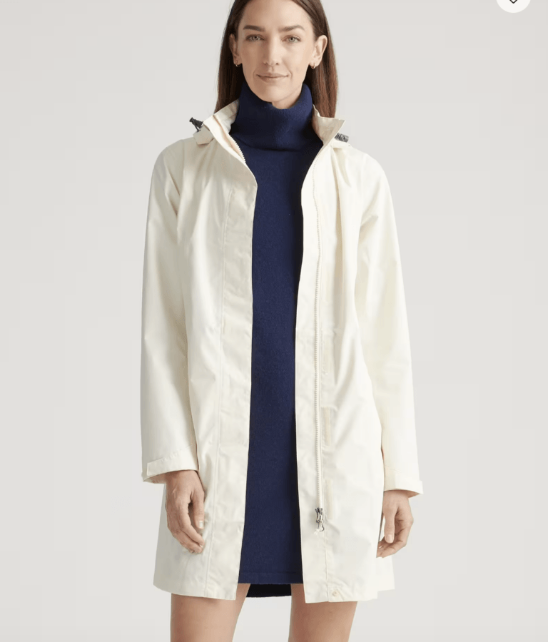 What to Wear for Derby 2026 - 9 A woman wearing a cream-colored, long-sleeve coat over a navy blue turtleneck dress stands facing forward against a plain background, embodying the elegance often seen in Kentucky Derby outfits.