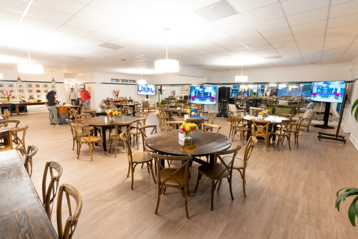 A Masters-Inspired Design Project You Need to See - 2 Spacious event room with round wooden tables and chairs, floral centerpieces, large TVs, and a few people near a display wall.