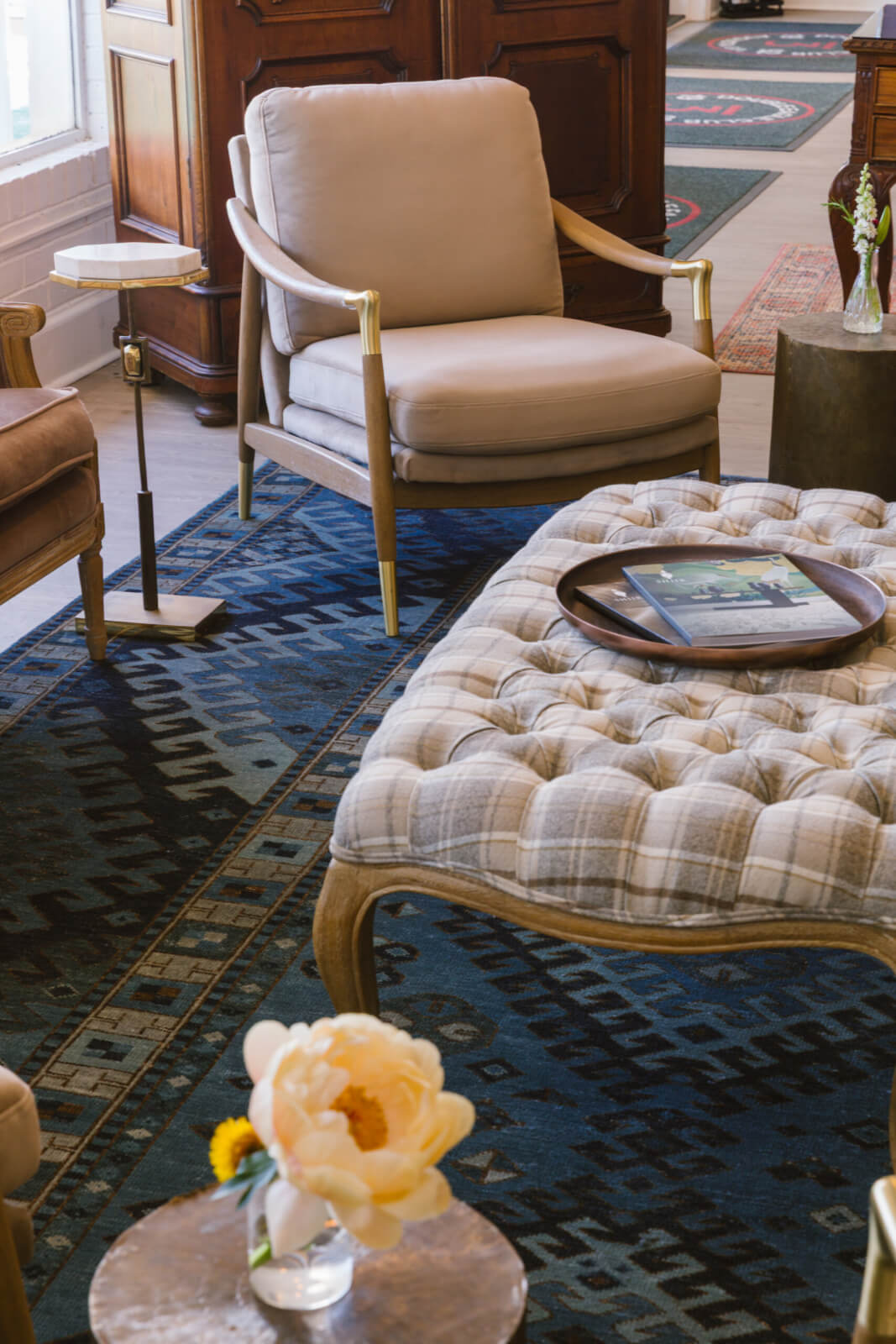 A Masters-Inspired Design Project You Need to See - 5 A beige armchair and a tufted ottoman with a tray and book sit on a patterned rug, showcasing a Masters-Inspired Design; a small table with a glass vase and flower is in the foreground.