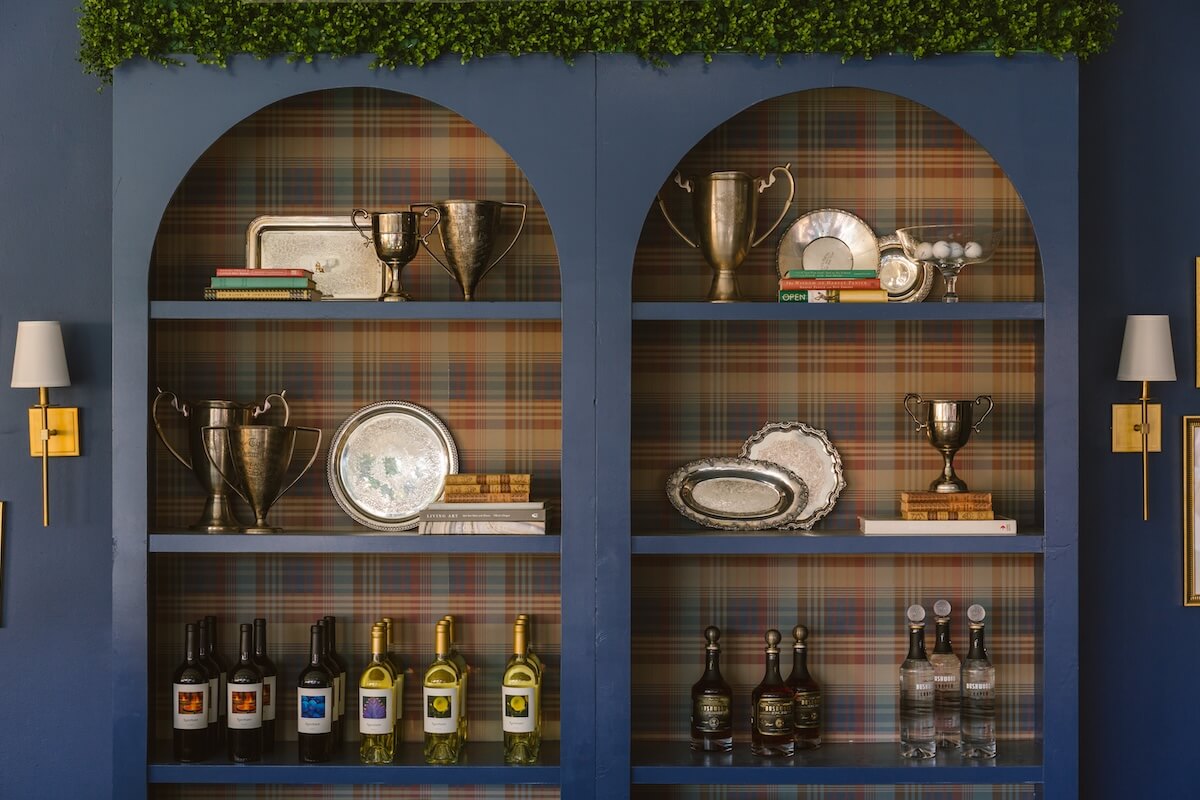 Two blue arched shelves showcase silver trophies, trays, stacked books, five wine bottles, and four bottles of clear liquor against a plaid-patterned back panel with overhead greenery—a true Masters-Inspired Design project.