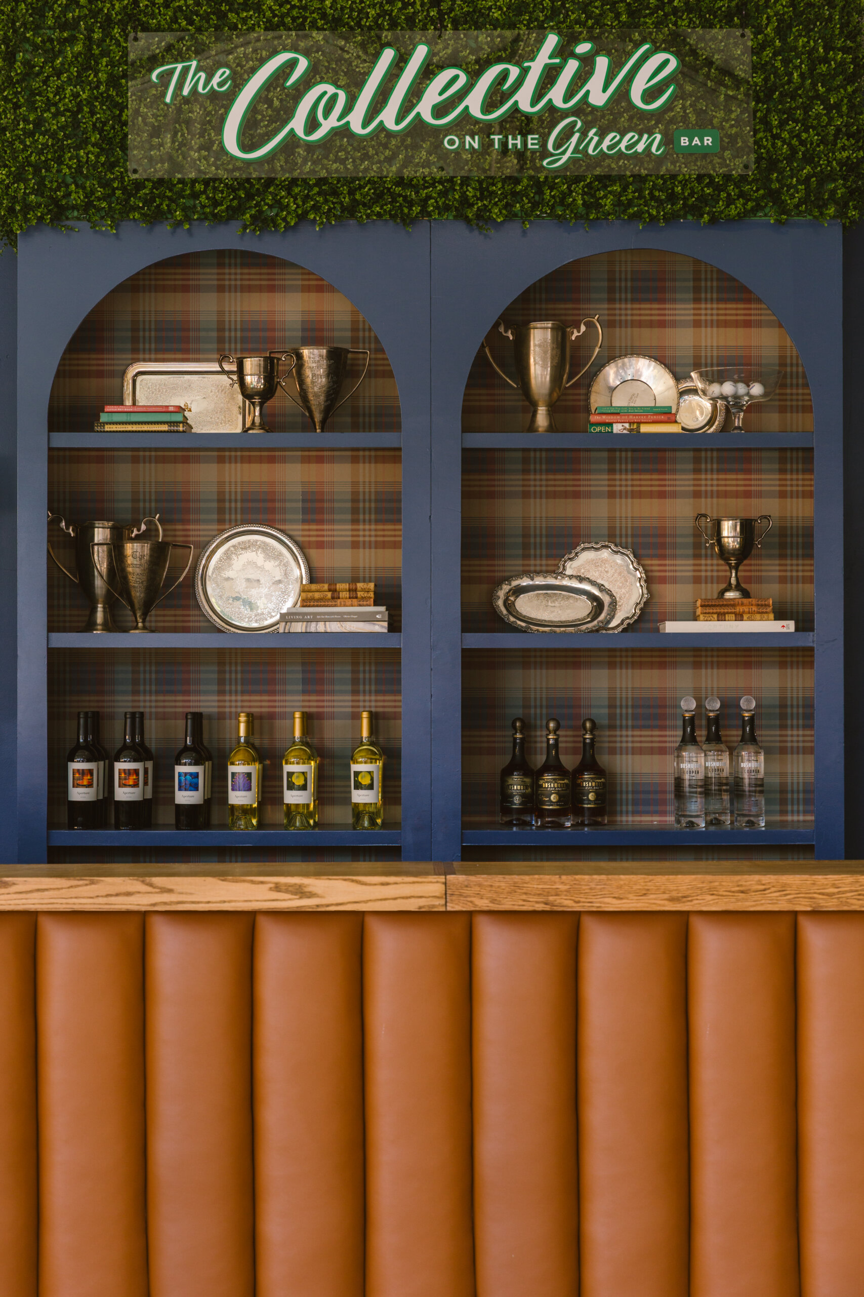 A Masters-Inspired Design Project You Need to See - 11 A bar shelf with silver trophies, trays, liquor bottles, and books rests beneath a green sign reading "The Collective on the Green Bar."