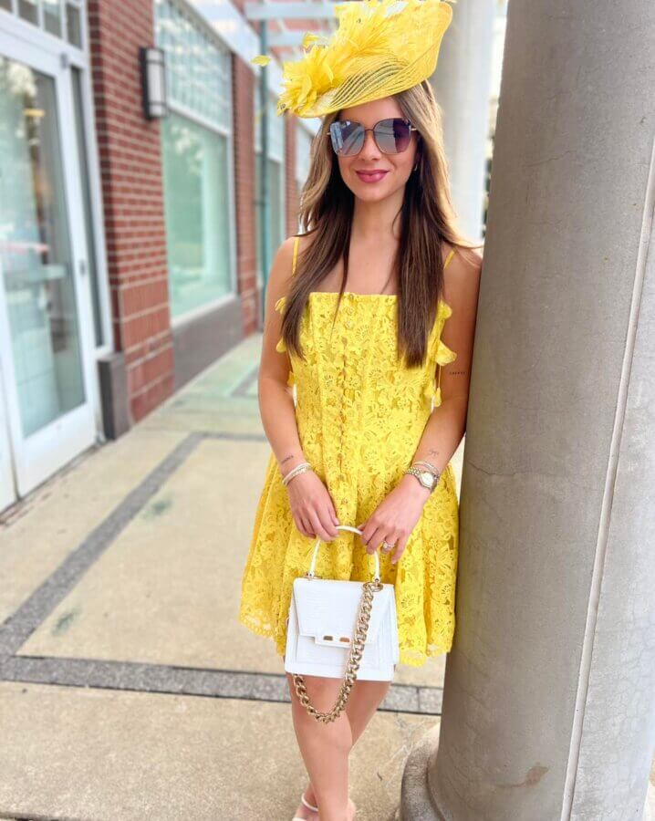 What to Wear for Derby 2026 - 2 Woman in a yellow lace dress and matching hat, holding a white handbag, stands by a column on a city sidewalk, embodying the elegance of classic Kentucky Derby outfits.