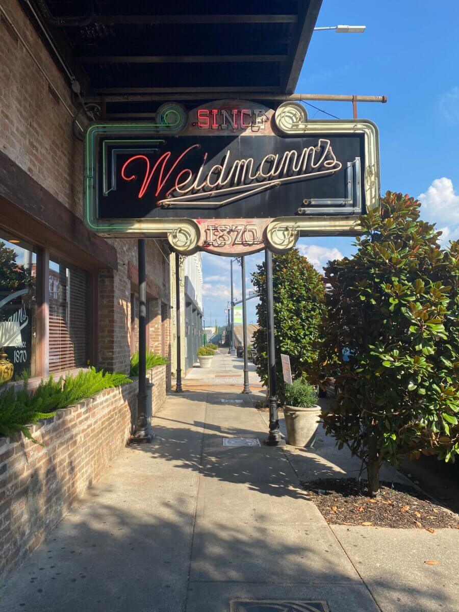 The South's Oldest Restaurants, Still Operating Today - 8 A vintage neon sign reading "Weidmann's Since 1870" hangs above a sidewalk outside a brick building on a sunny day—one of the South’s oldest restaurants still operating.