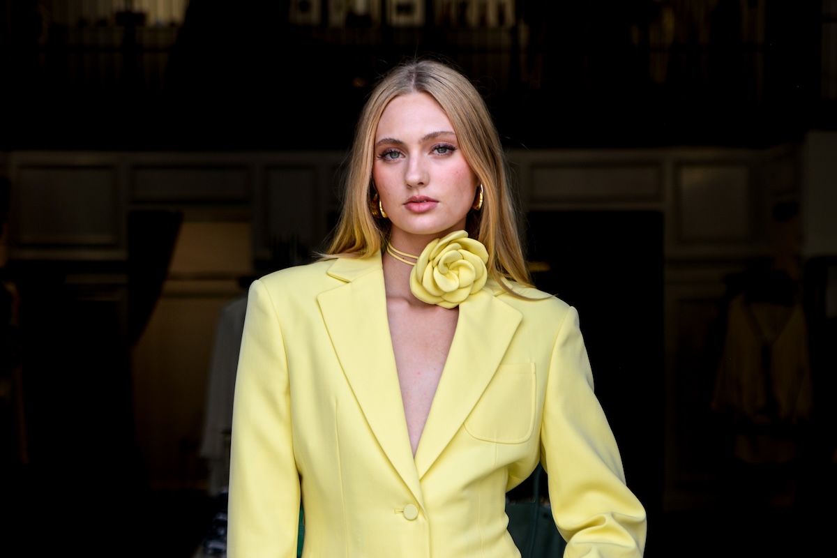 A woman with long blonde hair wears a yellow blazer and a large yellow flower choker, standing indoors with a dark background—an elegant look inspired by Nashville Boutiques.