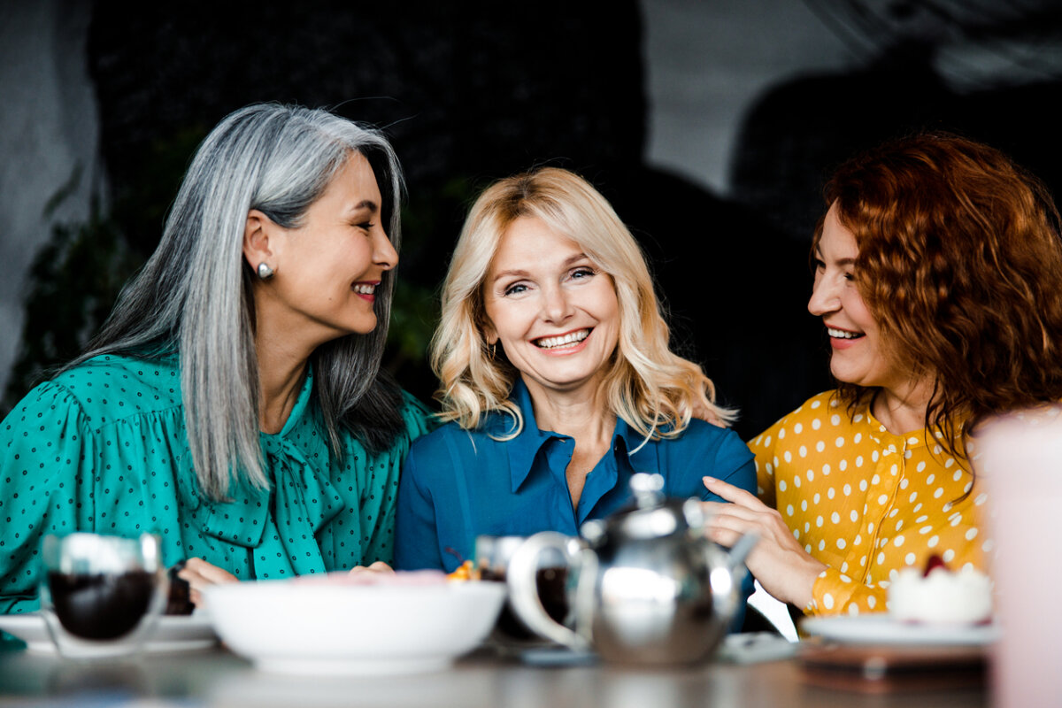 Three women sit at a table with tea and desserts, smiling and enjoying a conversation together, one of them radiantly sharing her recent deep plane facelift experience.