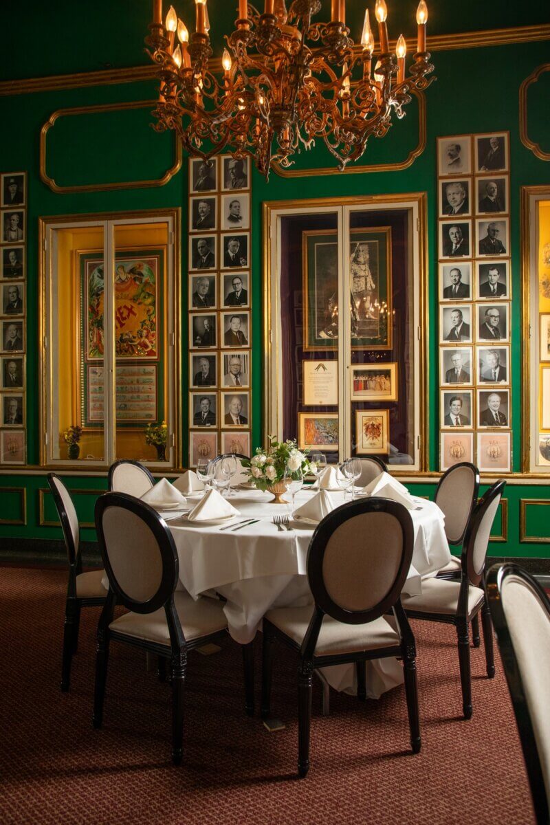 The South's Oldest Restaurants, Still Operating Today - 7 A round dining table with white tablecloth and chairs is set in an elegant room with green walls, gold accents, framed photos, and a chandelier—reflecting the timeless charm of one of the South’s oldest restaurants still operating.
