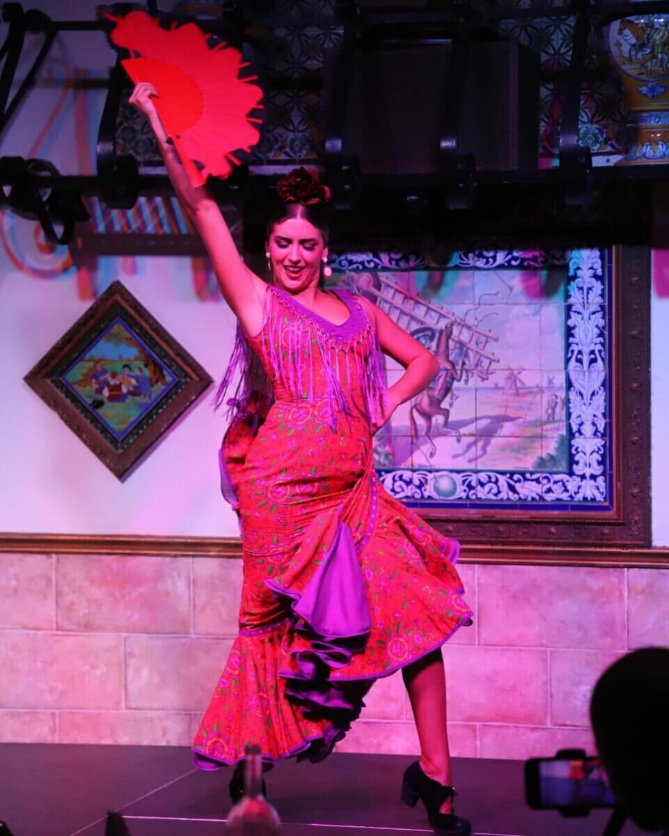 The South's Oldest Restaurants, Still Operating Today - 4 A flamenco dancer in a vibrant red and purple dress performs on stage, holding a red fan above her head, with colorful artwork and tiles in the background of one of the South’s oldest restaurants still in operation.