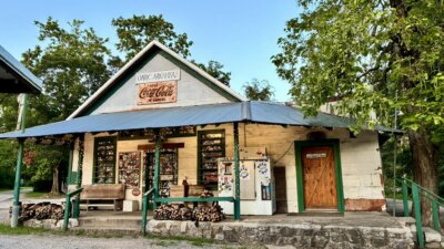 A rustic country store with a tin roof, woodpile, and vintage Coca-Cola sign stands among trees and greenery under a clear sky—reminiscent of the oldest restaurants in the South still in operation.