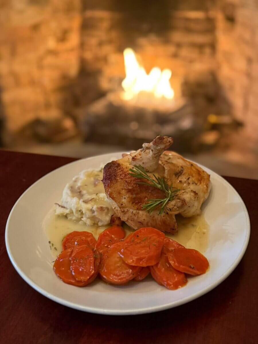 The South's Oldest Restaurants, Still Operating Today - 6 A plate of roast chicken with rosemary, sliced cooked carrots, and mashed potatoes sits on a table in front of a lit fireplace at one of the oldest restaurants in the South still operating today.