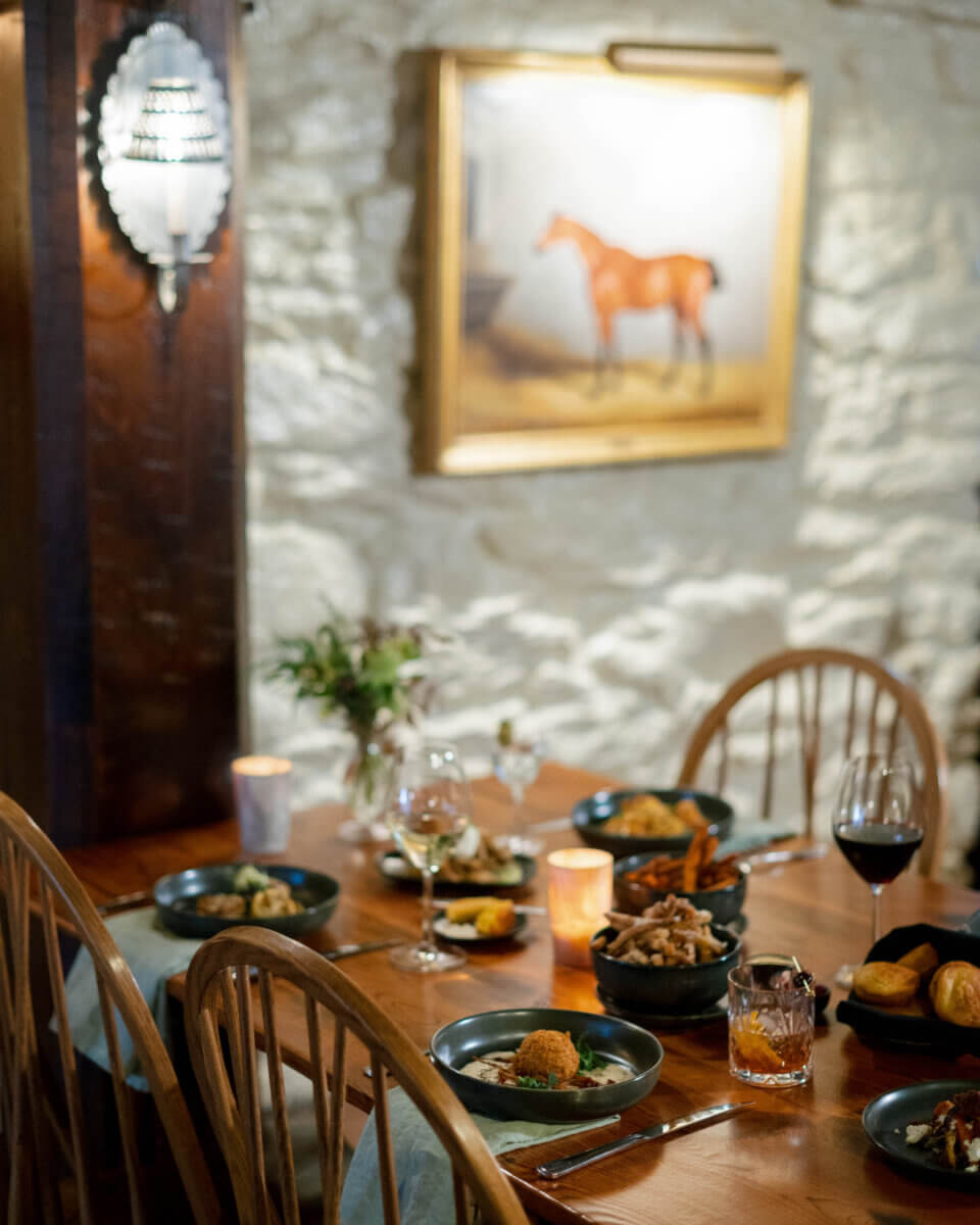 The South's Oldest Restaurants, Still Operating Today - 12 A wooden dining table set with plates of food, drinks, and candles in a warmly lit room evokes the charm of the South’s oldest restaurants still operating, with a horse painting on a stone wall in the background.