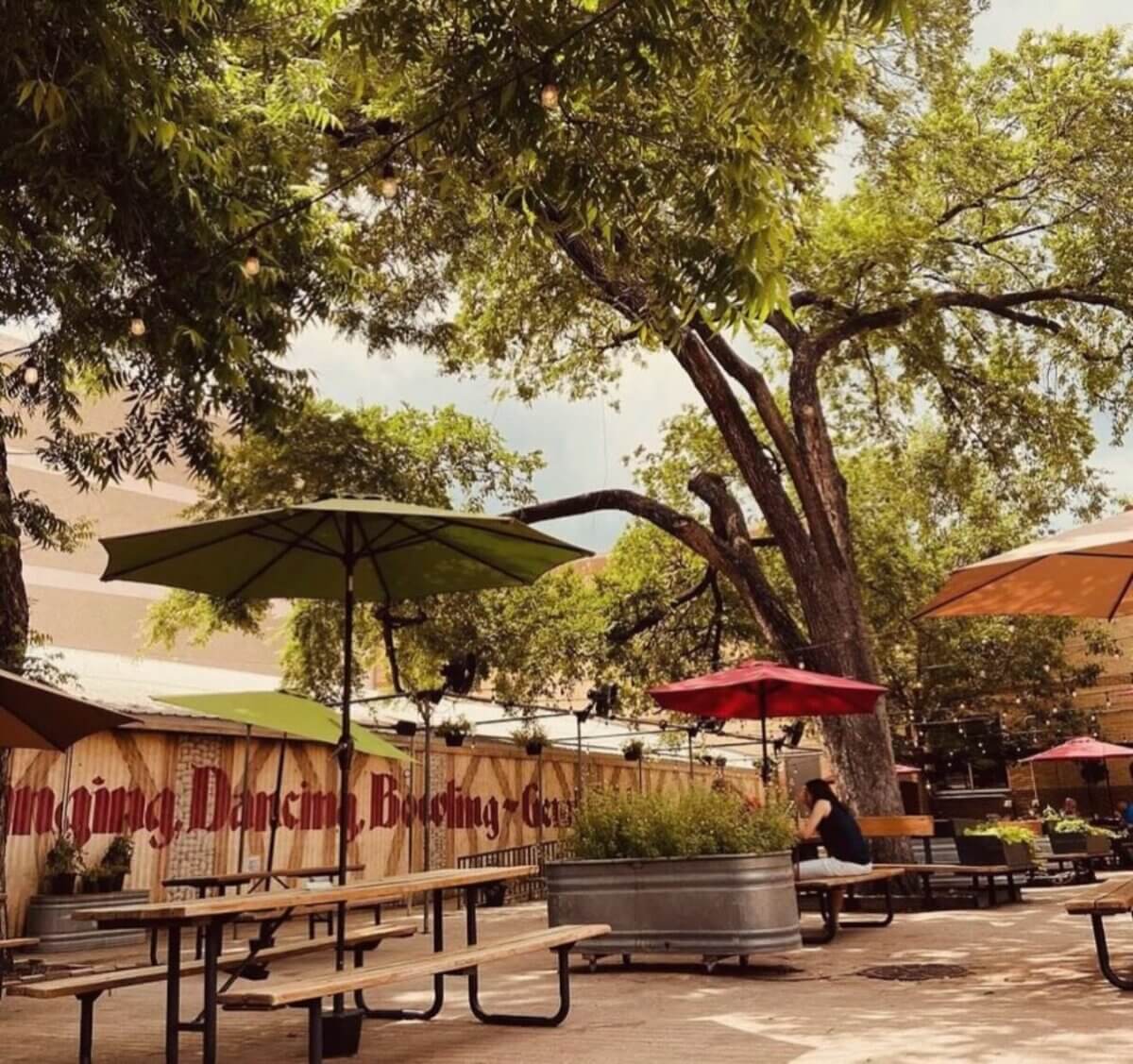 The South's Oldest Restaurants, Still Operating Today - 11 Outdoor seating area with picnic tables, large umbrellas, and shade from trees at one of the South's oldest restaurants still operating. A person sits alone at one table; a wooden fence with mural text is visible in the background.