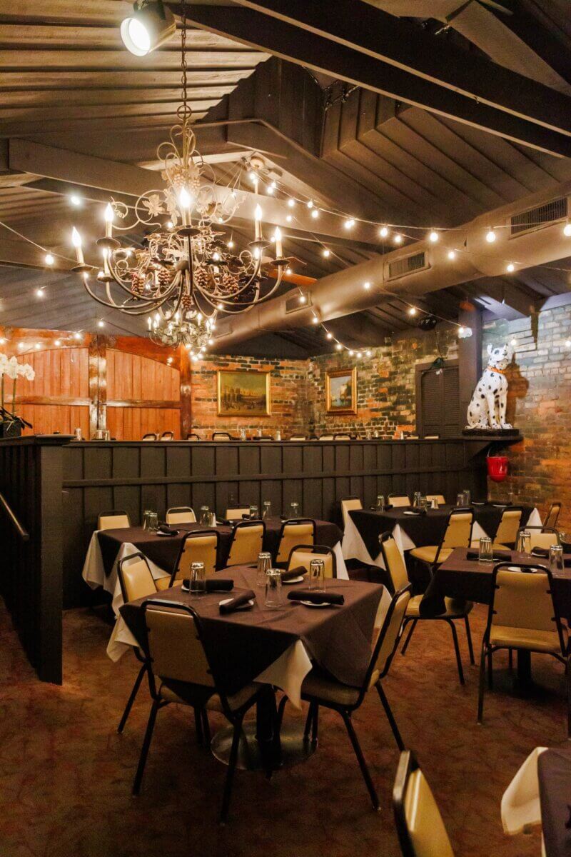 The South's Oldest Restaurants, Still Operating Today - 9 A dimly lit restaurant dining room in the South with round tables covered in white and brown tablecloths, a chandelier, string lights, and a large Dalmatian statue on a ledge—one of the oldest restaurants still operating.