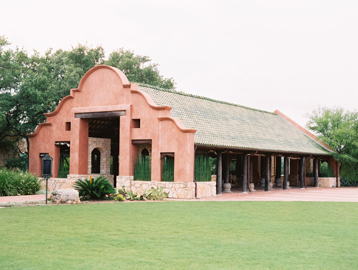 She's Behind Camp Lucy, a Dreamy Austin-Area Escape - 4 Single-story adobe-style pavilion with arched entrance, tile roof, and open sides, surrounded by grass, shrubs, and trees at Camp Lucy.