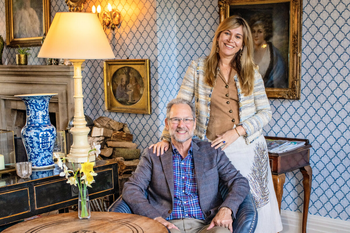 She's Behind Camp Lucy, a Dreamy Austin-Area Escape - 2 A smiling man sits in a chair while a woman stands behind him with her hand on his shoulder. They are in a traditionally decorated room at Camp Lucy, with framed portraits and patterned wallpaper.