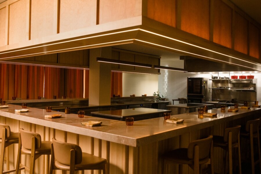 Modern restaurant interior with a square marble counter, high chairs, and carefully set glasses and napkins at each seat; open kitchen area visible in the background of this James Beard Award contender.