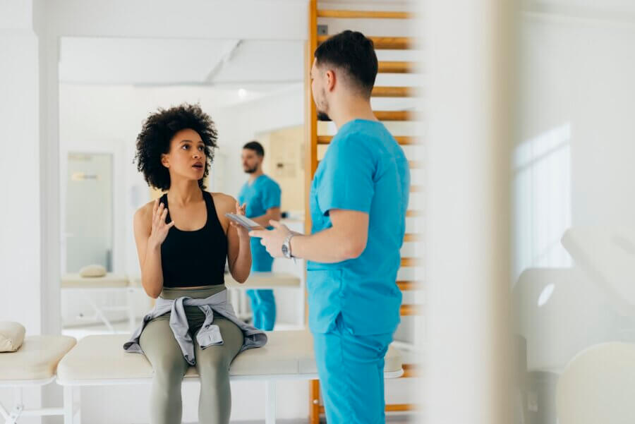 What the HECK is NAD? (And Should You Be Taking It?) - 2 A woman sits on an exam table in a medical clinic, talking to a healthcare professional in blue scrubs who is holding a clipboard and discussing the potential benefits of NAD supplements.