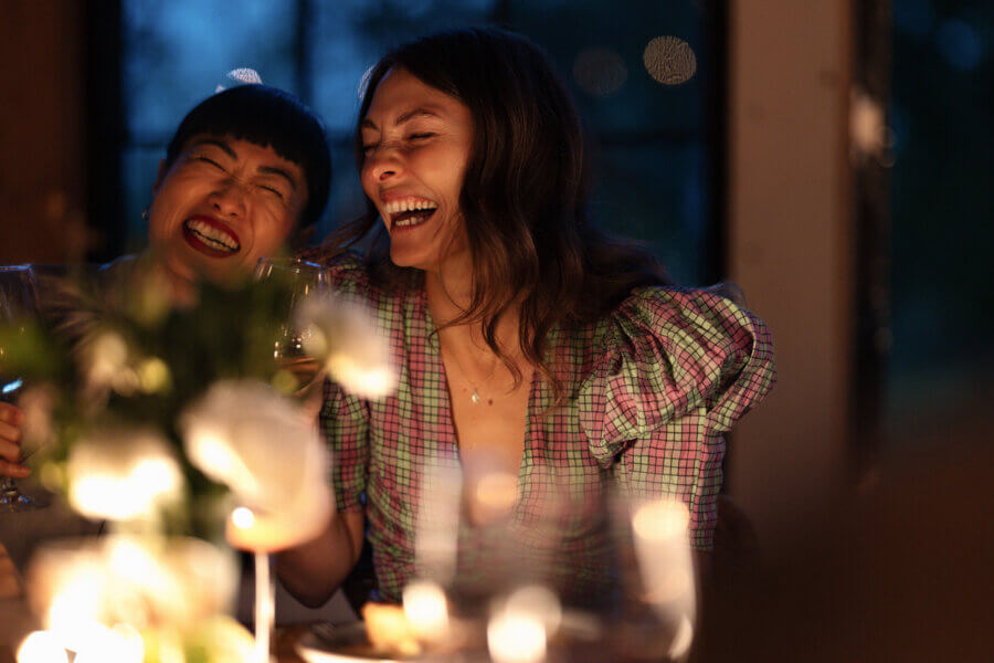 Two women sit close together at a dinner table, smiling and laughing as they share personal finance stories, with soft lighting and blurred flowers in the foreground.