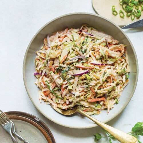 A bowl of coleslaw with shredded cabbage, carrots, and red onions on a light surface next to a cutting board with chopped green onions and a plate with forks.
