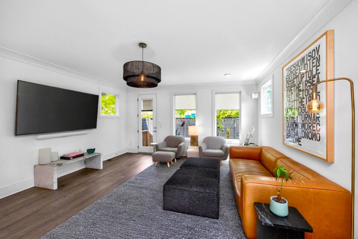 Modern living room on Eastmoreland St, Nashville, featuring a wall-mounted TV, tan leather sofa, two gray chairs, large framed artwork, and a floor lamp on a dark rug with wood flooring and white walls. Welcome to 217B.