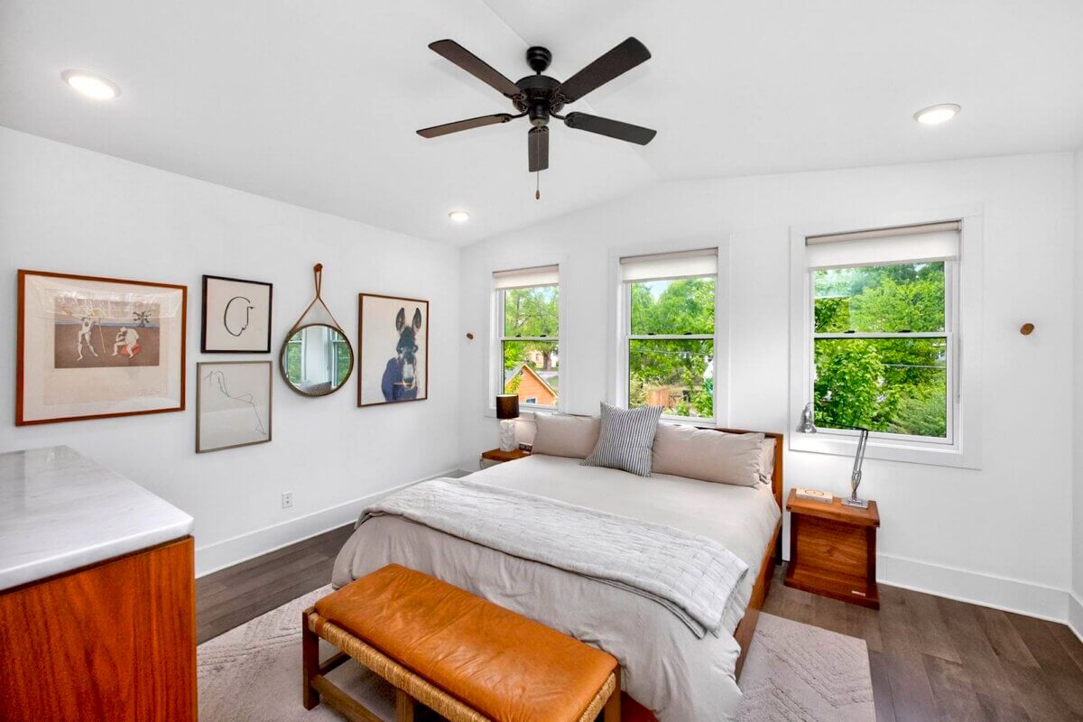 Modern bedroom in Eastmoreland St Nashville with a large bed, wooden bench, dresser, wall art, and three windows letting in natural light. Ceiling fan and minimalist decor complete this bright, clean Nashville TN space.