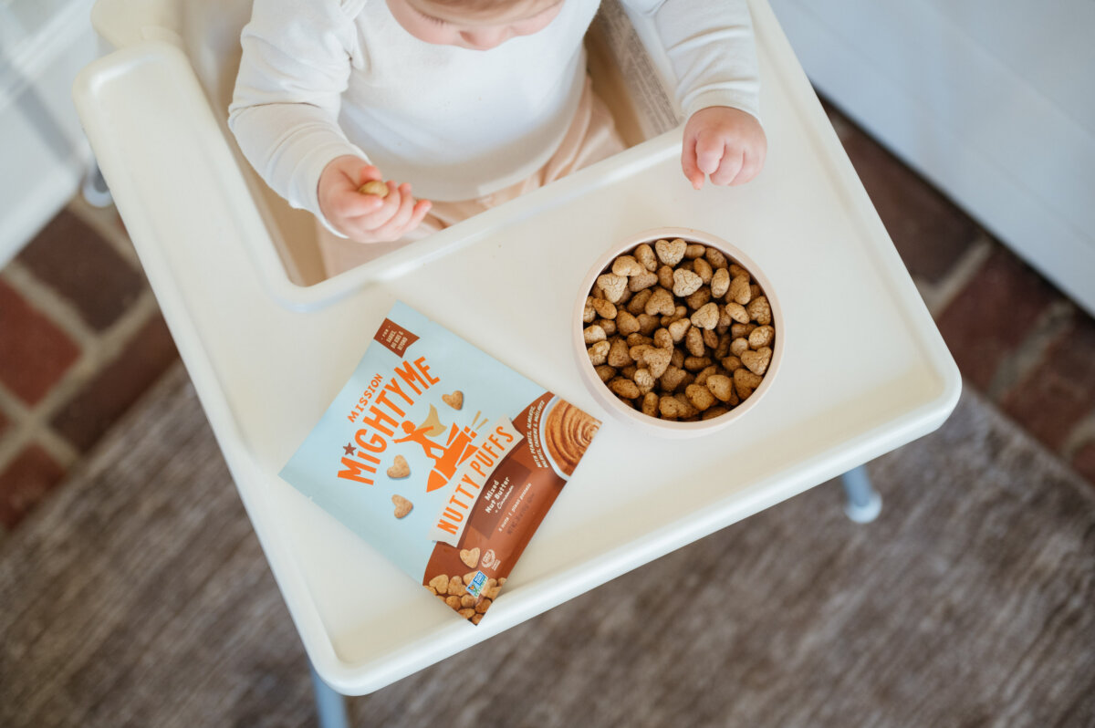 This Mom is on a Mission to Change the Future of Food Allergies - 3 A baby sits in a high chair, reaching for heart-shaped snacks in a bowl on the tray, with a blue "Mighty Me Nutty Puffs" package beside it—supporting the mission to tackle the food allergy epidemic.