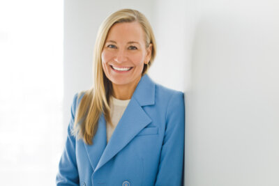 Catherine Jaxon, co-founder of Mission MightyMe, with long blonde hair, wearing a light blue blazer and white top, stands smiling against a white wall.