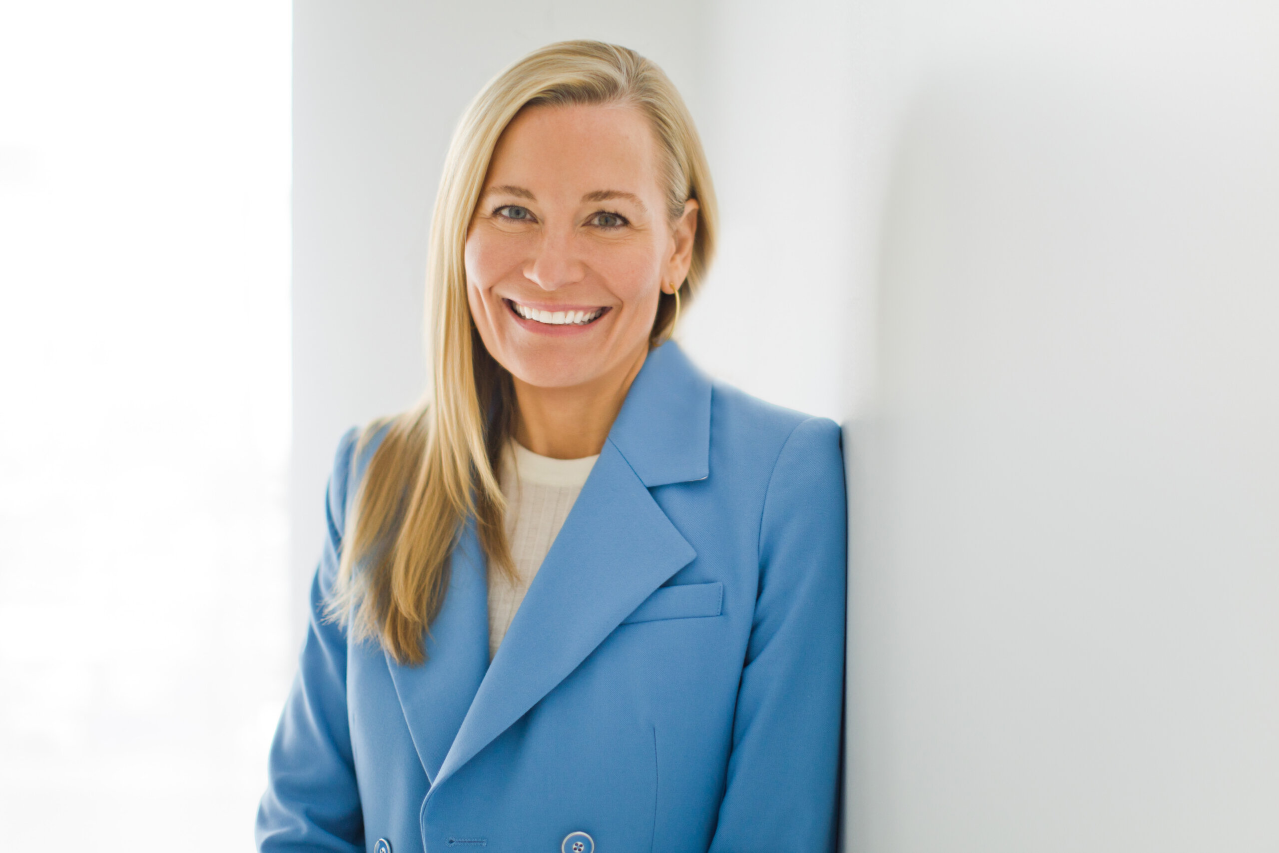 Catherine Jaxon, co-founder of Mission MightyMe, with long blonde hair, wearing a light blue blazer and white top, stands smiling against a white wall.