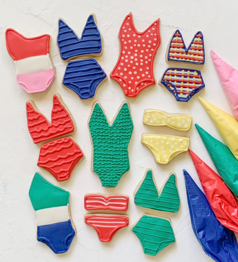 Meet Sally McKay, the Founder of Sally's Confections - 7 Assorted cookies, baked with love and decorated to look like colorful swimsuits and bikinis, are arranged on a white surface next to several piping bags filled with icing.