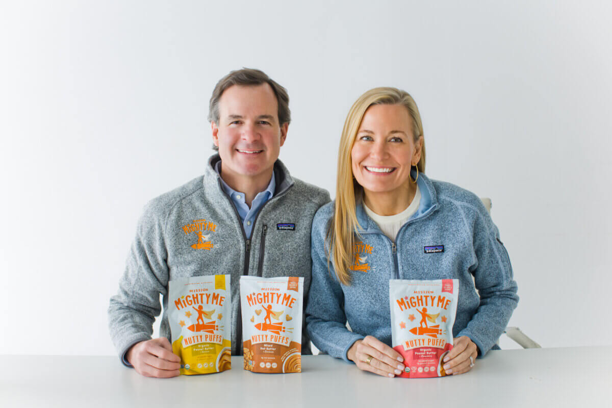 This Mom is on a Mission to Change the Future of Food Allergies - 2 A man and a woman in matching gray jackets sit at a table, each holding a bag of Mighty Me Nutty Puffs snacks, highlighting the mission to tackle the food allergy epidemic.