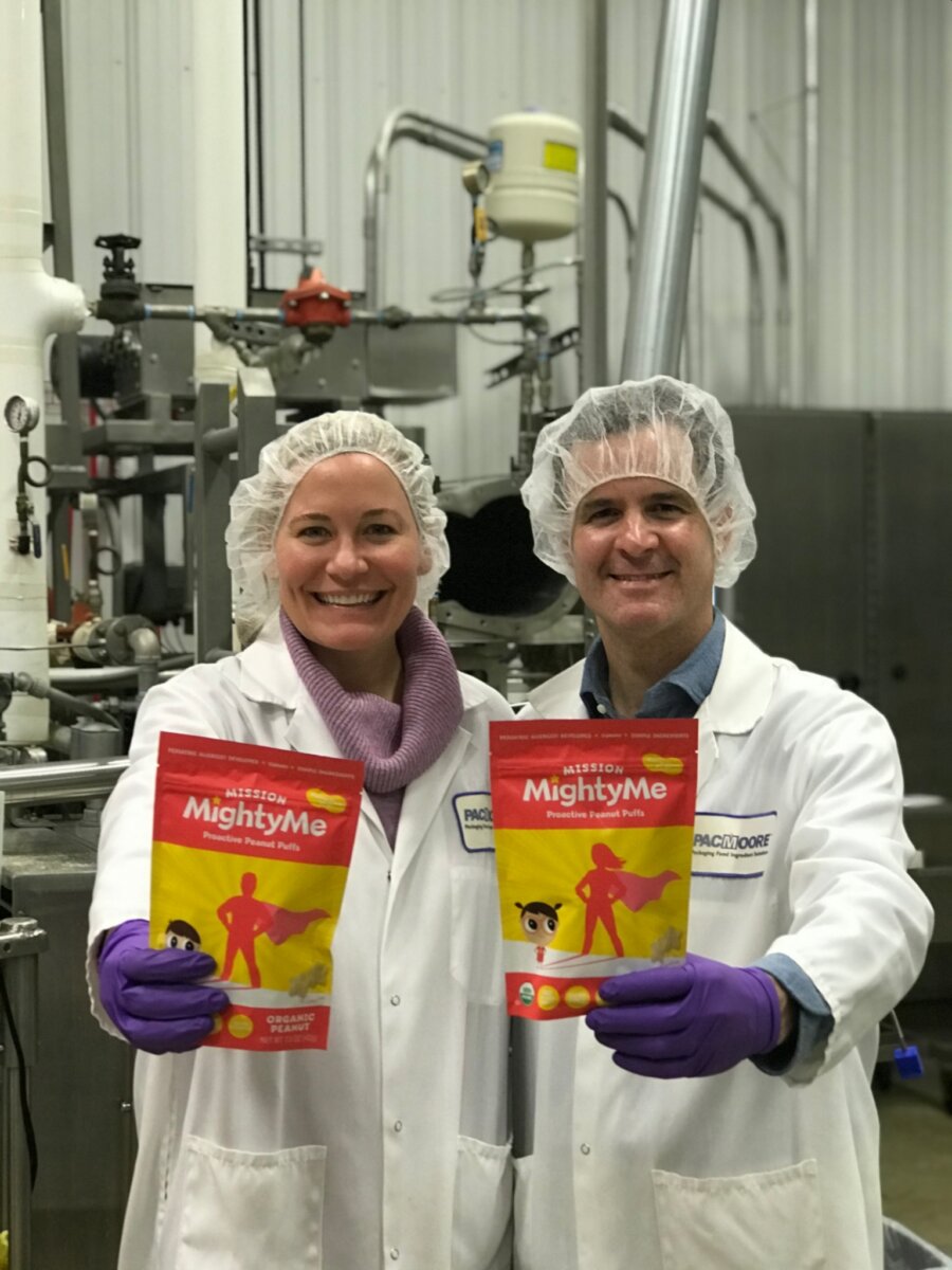 This Mom is on a Mission to Change the Future of Food Allergies - 6 Two people in lab coats and hairnets stand in a factory setting, each holding a red and yellow package labeled "MightyMe."