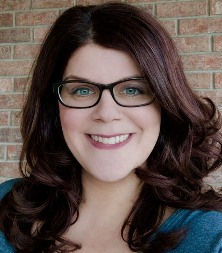 In Memoriam: Jessica Spinozzi Posel's Guide to Carefree, Arizona - 2 A woman with wavy dark brown hair and glasses smiles at the camera, standing in front of a brick wall, capturing the relaxed vibe of an Arizona Getaway.