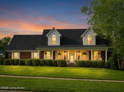 Charming two-story brick house at 7210 Rambo Way, Louisville KY, featuring a large front porch, dormer windows, and trimmed shrubs, beautifully photographed at dusk with the interior lights glowing.