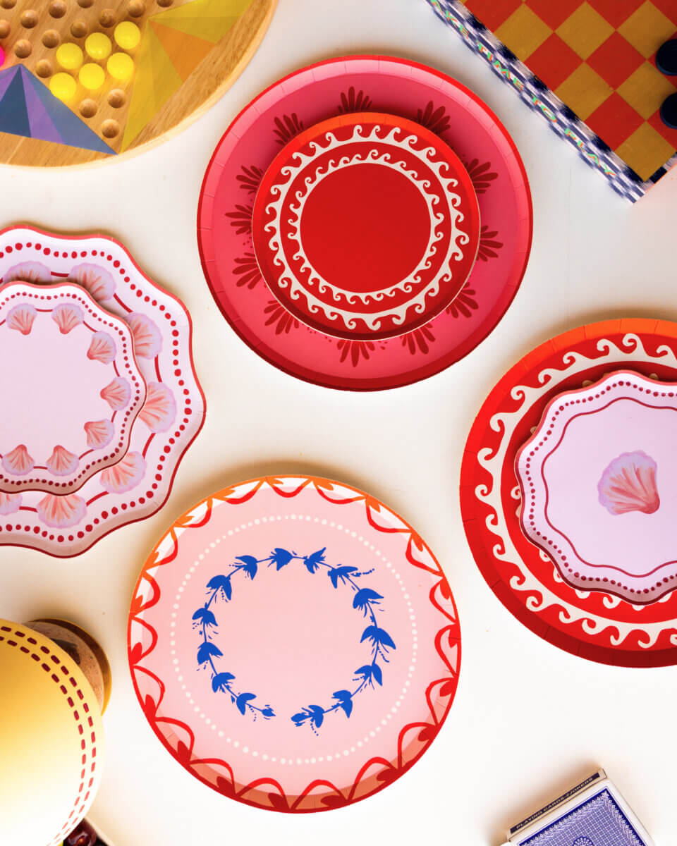 Table Two Studio's Luxe Paper Plates Will Change How You Entertain - 3 Four luxury paper plates by Table Two Studio, featuring red, pink, and blue patterns, sit on a white surface alongside board game pieces and other objects.