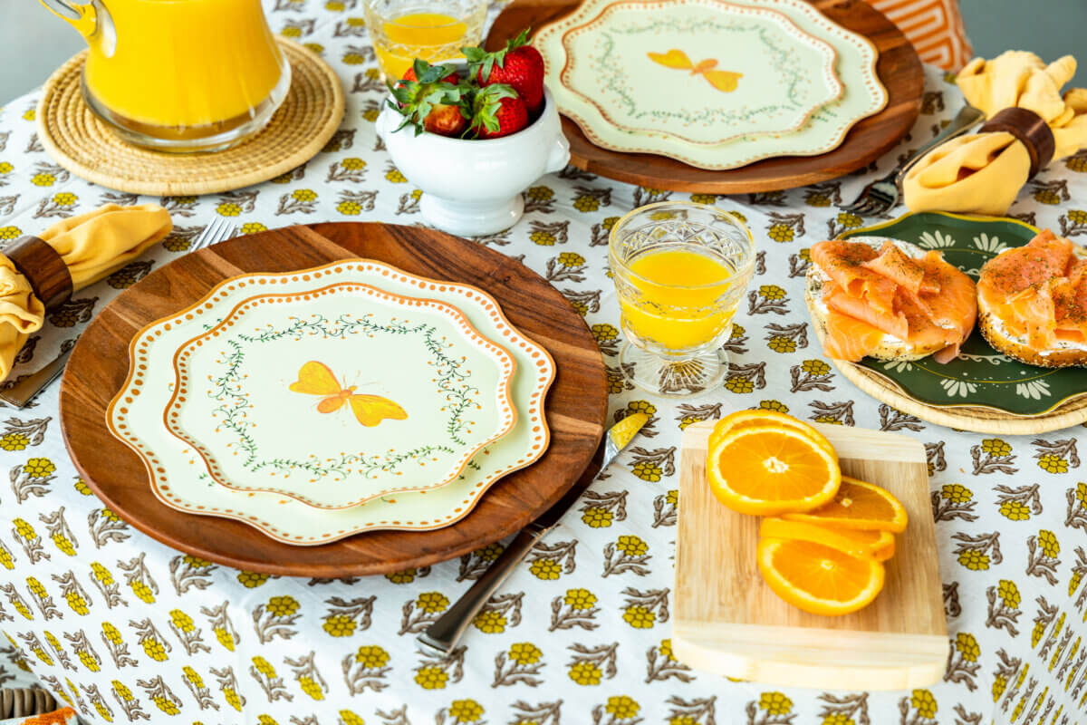 Table Two Studio's Luxe Paper Plates Will Change How You Entertain - 9 A brunch spread on a yellow floral tablecloth, featuring orange juice, bagels with lox, and fruit.