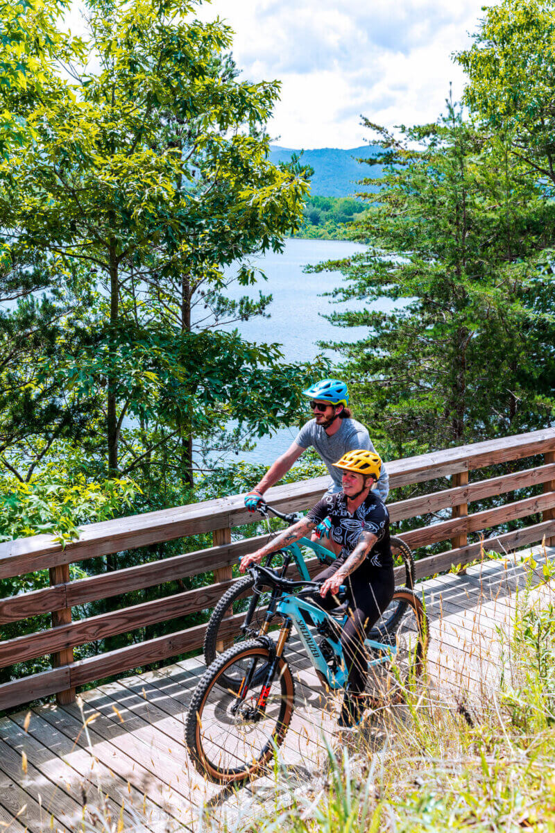 4 Summer Weekend Trips from Atlanta - 9 Two people wearing helmets ride bicycles on a wooden bridge surrounded by trees, with a lake and hills visible in the background—perfect for Summer Weekend Trips near Atlanta.