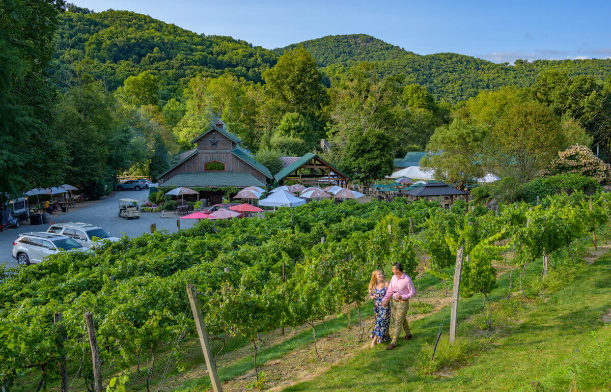 4 Summer Weekend Trips from Atlanta - 7 Two people walk through a vineyard with a rustic building, outdoor seating, parked cars, and green hills in the background under a blue sky—an ideal spot for relaxing summer weekend trips from Atlanta.