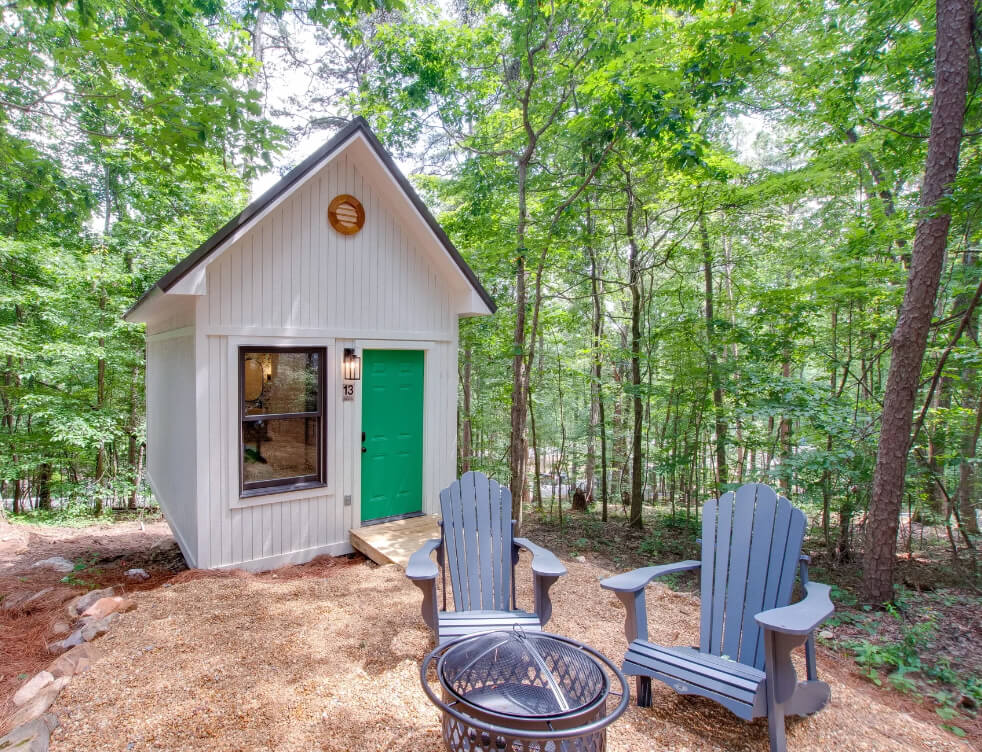 4 Summer Weekend Trips from Atlanta - 5 A small white cabin with a green door is surrounded by trees—perfect for summer weekend trips from Atlanta—with two Adirondack chairs and a fire pit set on gravel in front.