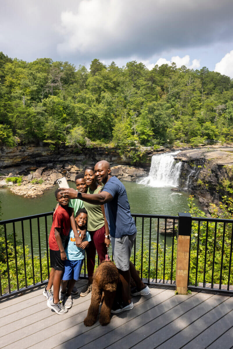 4 Summer Weekend Trips from Atlanta - 4 A family with three children and a dog stands on a wooden deck overlooking a waterfall and lush greenery, enjoying one of Atlanta's perfect summer weekend trips on a partly cloudy day.