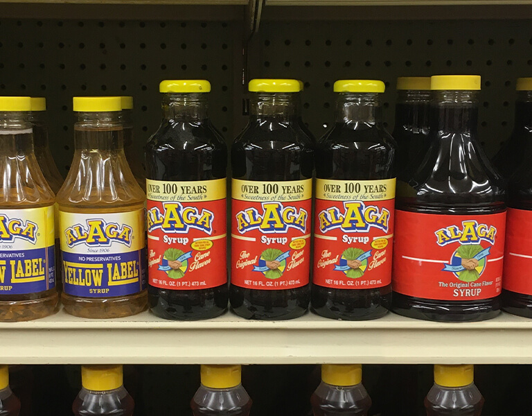ALAGA Syrup: Alabama's IYKYK Staple - 2 Bottles of ALAGA Syrup, an Alabama Staple, including Yellow Label and Original Cane, are lined up on a grocery store shelf.