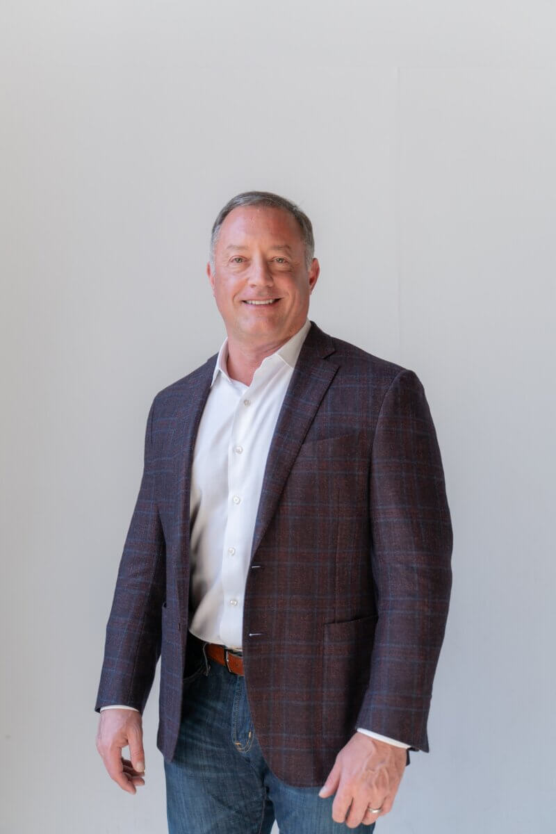 Is It Time for Hormone Therapy? What You Need to Know - 2 A man wearing a white shirt, checked blazer, and jeans stands against a plain light background, smiling at the camera.