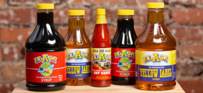 Five bottles of ALAGA Syrup, an Alabama staple, including three yellow label syrups and two red southern seasoned hot sauces, are arranged in a row on a wooden surface in front of a brick wall.
