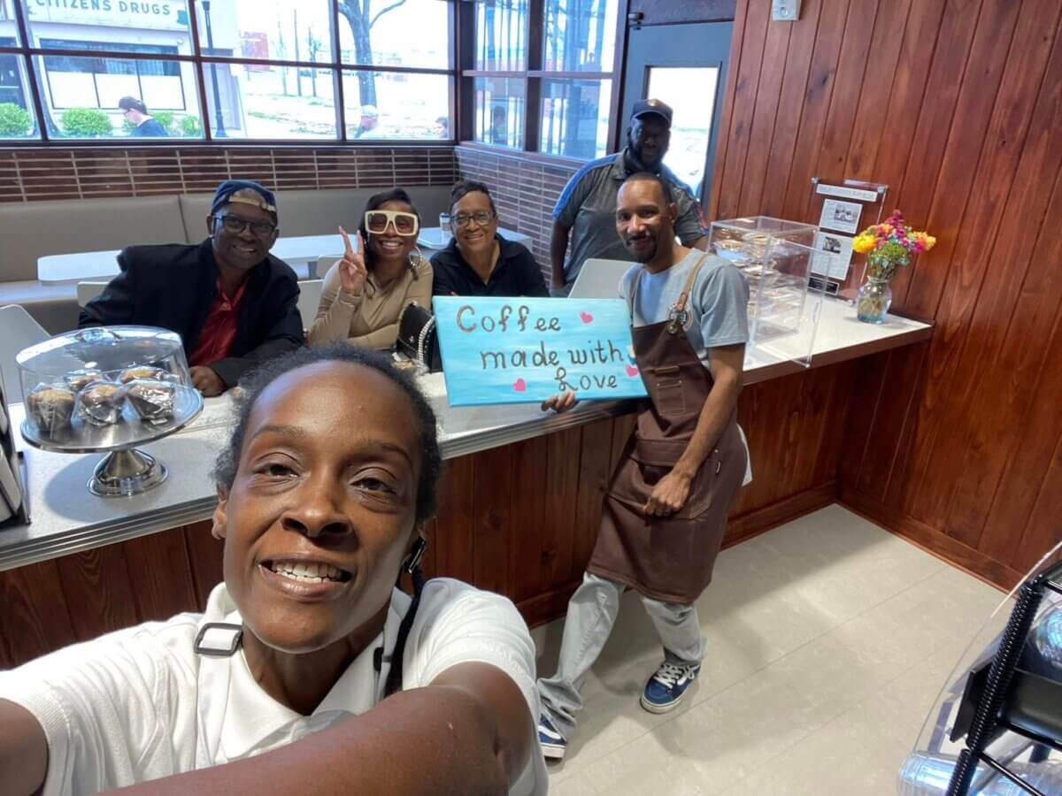 How Two BHAM Creatives Built a Coffee Shop Centered on Connection - 4 A group of six people smile behind a cafe counter; one holds a sign that reads "Coffee made with love." Cupcakes and flowers are displayed on the counter.
