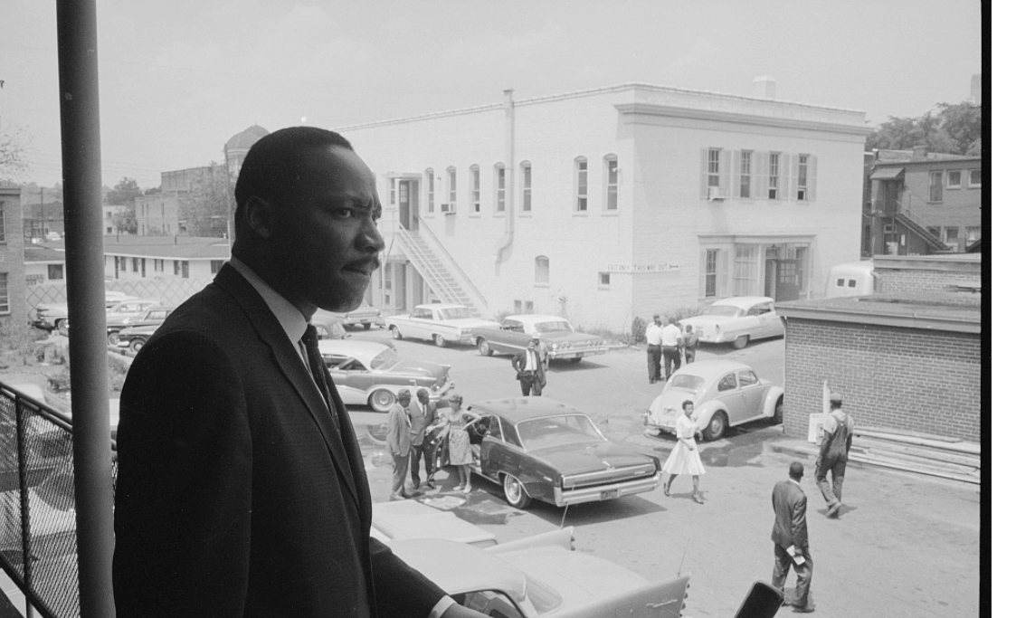 How Two BHAM Creatives Built a Coffee Shop Centered on Connection - 6 Martin Luther King, Jr. in a suit stands on a balcony at the A.G. Gaston Motel overlooking a parking lot with several parked cars and people walking below near a white building.