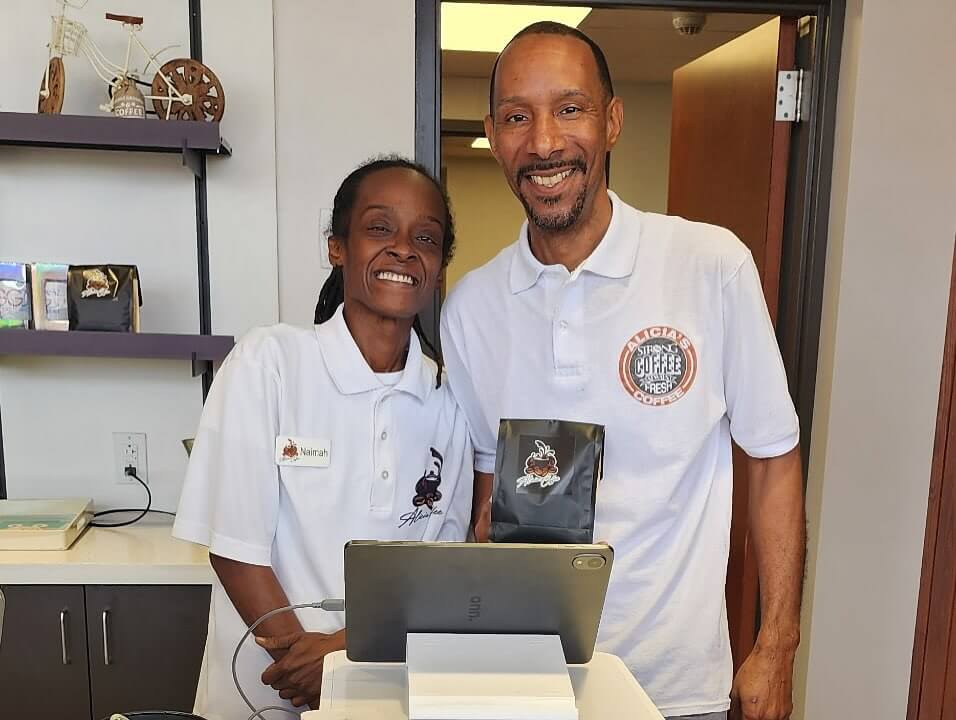 How Two BHAM Creatives Built a Coffee Shop Centered on Connection - 3 Two people in matching white shirts stand behind a counter, smiling. One holds a bag of coffee, and a tablet register is visible in front of them.