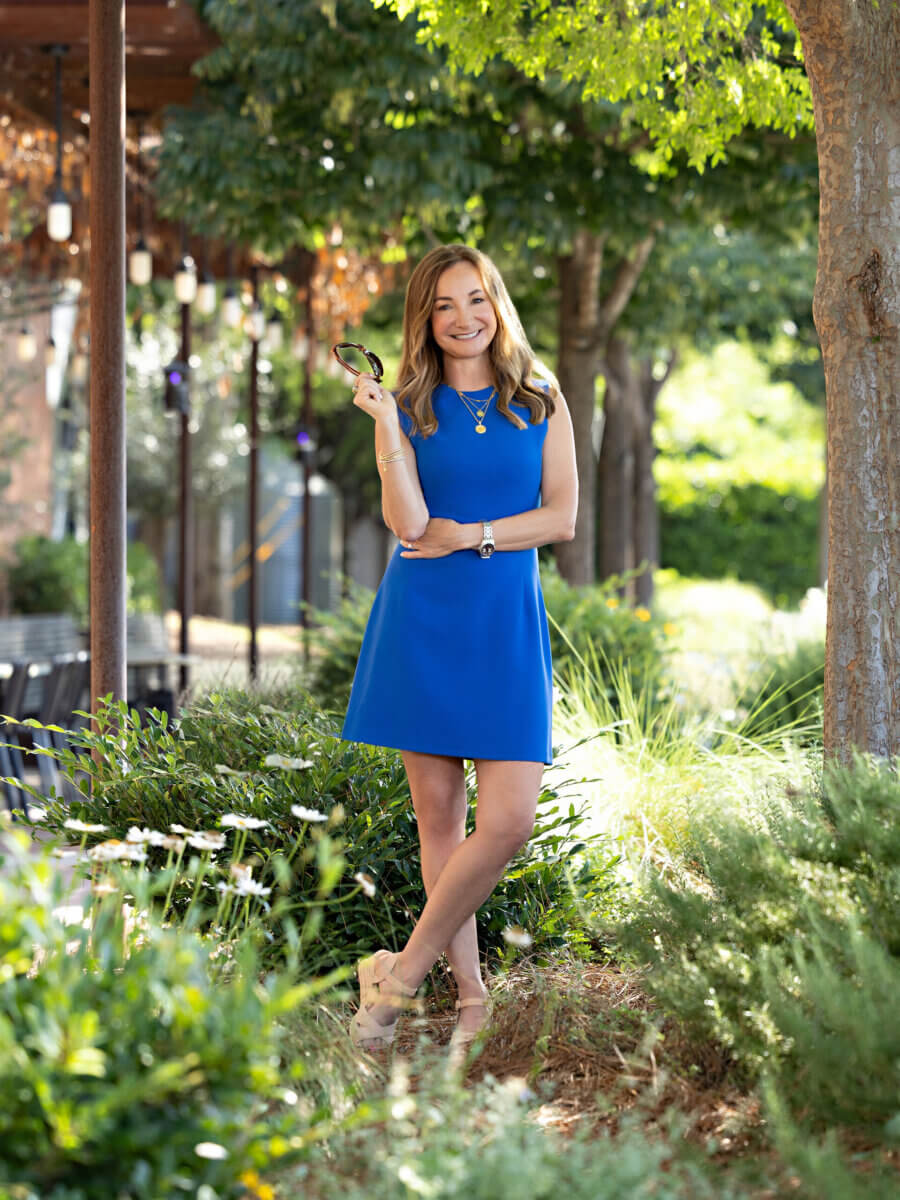 Meet the PR Pro Behind Some of Luxury's Biggest Brands - 4 A woman in a blue dress stands outdoors, holding sunglasses and smiling.