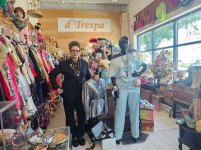 LaRoyce Marsh stands inside d'Trespa, a Birmingham vintage boutique, surrounded by colorful garments, hats, accessories, and two mannequins in retro outfits.