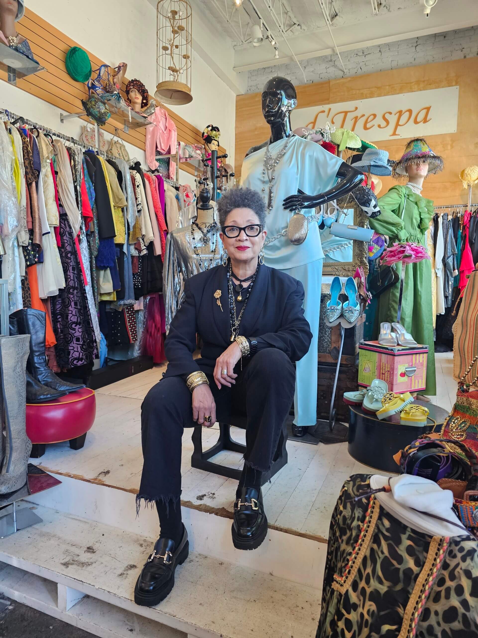 She Curates One of Birmingham’s Top Vintage & Consignment Boutiques - 2 LaRoyce Marsh wearing black clothing and glasses sits on a chair in a Birmingham vintage consignment boutique, surrounded by racks of colorful clothes, mannequins, and accessories.